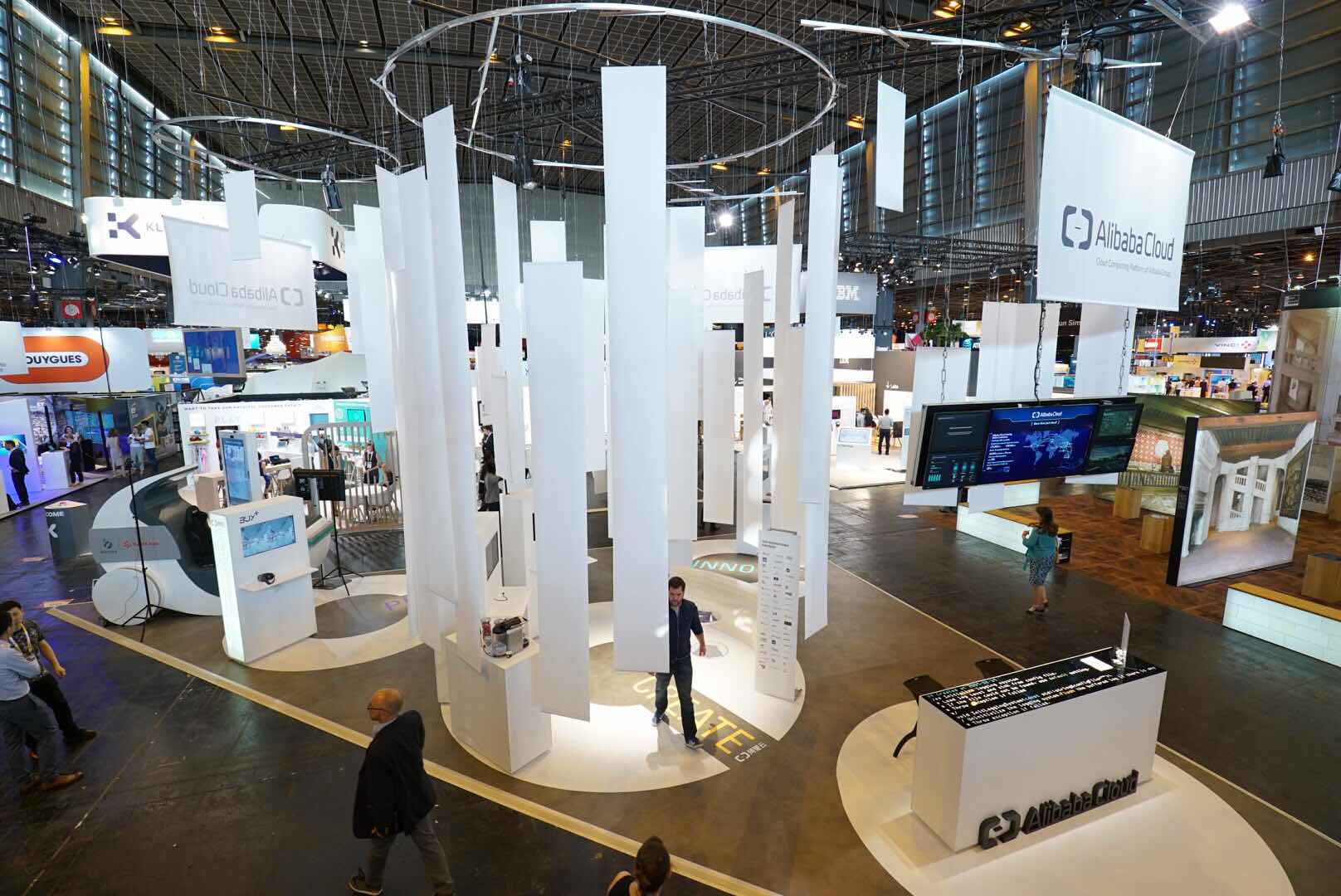  A large exhibition space with many booths, people walking around, and hanging signs, one of which says Alibaba Cloud.